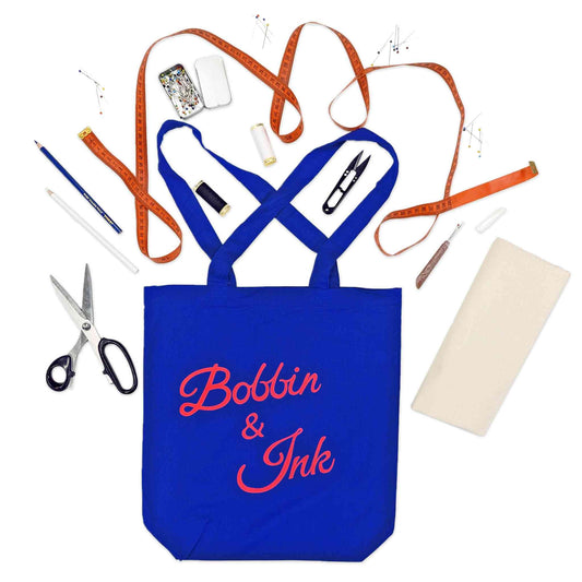 Blue tote bag with 'Bobbin & Ink' text, surrounded by sewing items on a white background